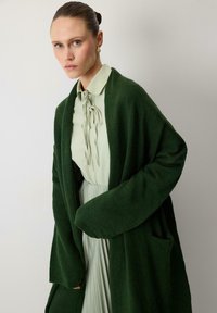 Long green knit cardigan with a loose fit, pocket detail, and ribbed cuffs, worn over a pale green, tied-neck blouse and pleated skirt.