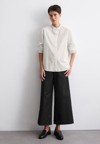 Chemise blanche à manches longues avec des pois noirs, col rond, fermeture à boutons à l'avant ; associée à un pantalon large noir et des chaussures noires à enfiler.