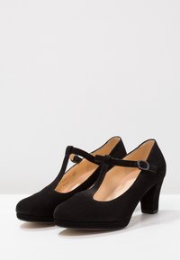 Tacones T-bar de ante negro con punta redonda, correa ajustable en el tobillo y diseño de tacón grueso. Forro interior suave.