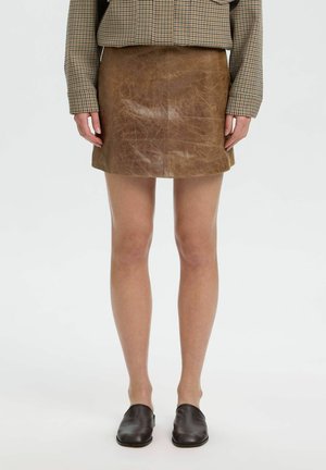 Femme portant une mini-jupe en cuir marron, une veste à carreaux et des chaussures noires sans lacets, debout devant un fond clair uni.