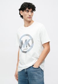 Camiseta de algodón blanca con un logo circular en azul y negro que incluye el texto "MK" y "MICHAEL KORS". Combinada con jeans de mezclilla azules.