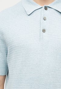 Ανοιχτό μπλε υφασμάτινο πόλο μπλουζάκι με κοντά μανίκια, τρία γκρι κουμπιά και επίπεδη πλεκτή γιακά, που φοράει ένα άτομο.