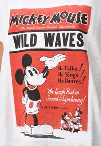 T-shirt blanc avec un graphique rouge et noir de Mickey Mouse accompagné du texte « WILD WAVES » et de détails de dessin animé. Le texte inclut des phrases promotionnelles.