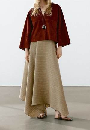 Femme portant un haut ample couleur rouille, une longue jupe asymétrique beige, un collier pendentif et des sandales plates marron, debout sur un sol gris.