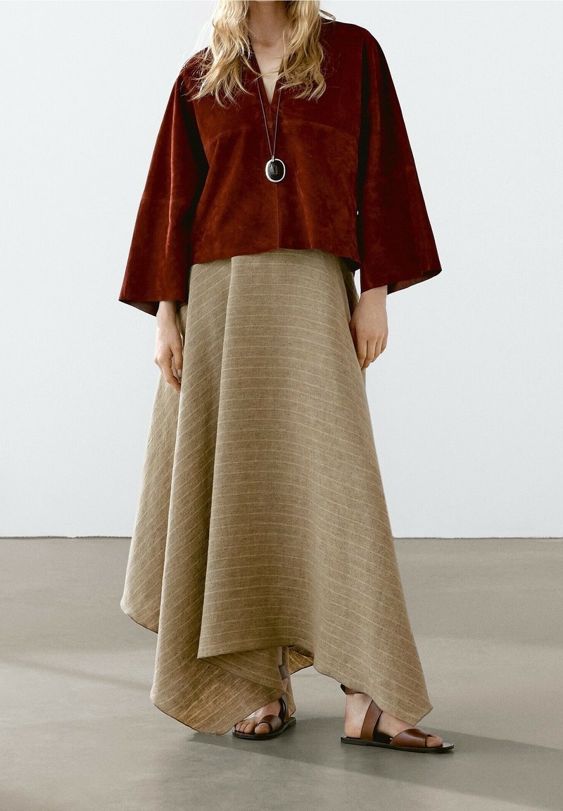 Femme portant un haut ample couleur rouille, une longue jupe asymétrique beige, un collier pendentif et des sandales plates marron, debout sur un sol gris.