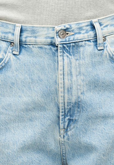 Φωτεινό γαλάζιο τζιν με μεταλλικό κουμπί και θηλιές για ζώνη, φορεμένο πάνω από τη μέση ενός γκρι ριγωτού πουκαμίσου.