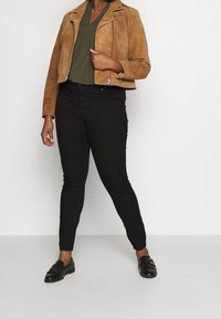 Ruskea mokkajakku moottoripyöräilyyn, mustat istuvat housut ja mustat loafers-kengät. Jakussa on vetoketjukiinnitys ja napitusyksityiskohtia.