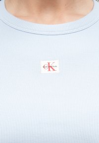 Jasnoniebieski, prążkowany top na szyję z białą metką z czerwonym logo "CK". Dekolt jest zaokrąglony i lekko teksturowany.