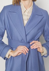 Abrigo de lavanda con un acabado texturizado, solapas anchas y un cinturón atado, superpuesto a una camisa crema a rayas con un cuello clásico.