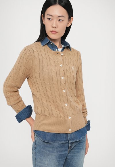 Bēšs adīts kardigans ar ribotiem apakšmalu un piecām pogām, valkāts virs zilas džinsa krekla. Piedāvā garas piedurknes ar salocītām aprocēm.