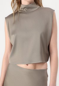 Ärmelloses Crop-Top aus weichem Taupe-Stoff mit hohem Kragen und glatter Textur. Verfügt über einen rohen Saum und subtile silberne Steppdetails.