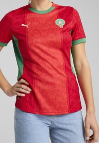 Osoba ubrana w marokański T-shirt piłkarski w kolorze czerwonym i zielonym z wzorami w gwiazdki i herbem, w połączeniu z jasnoniebieskimi dżinsami, stojąca z jedną ręką na biodrze.