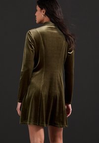 Zielona oliwkowa mini sukienka z weluru, z długimi rękawami i dopasowanym krojem, z rozkloszowanym dołem oraz subtelnym detalem szwów.