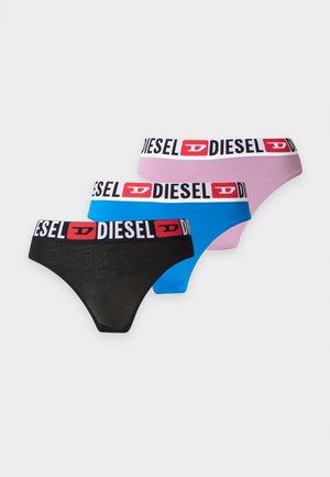 Frau steht barfuß, trägt ein gewaschenes schwarzes Croptop und rosa Unterwäsche der Marke Diesel vor einem einfachen weißen Hintergrund.