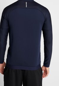 Marine langærmet sportstrøje med strukturerede skulderpaneler, blødt stof og tætsiddende design. Har logo på nakken bagpå.