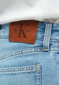 Világoskék farmernadrág barna bőr címkével, amelyen dombornyomott „Calvin Klein” logó és „CK” monogram látható a derékrész közelében.