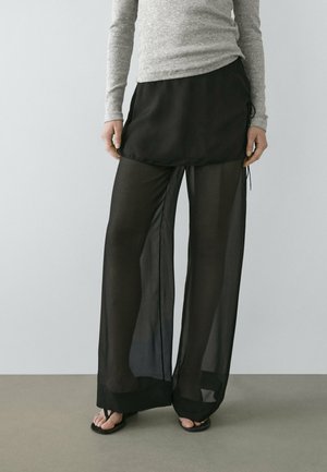 Persona che indossa pantaloni neri trasparenti a gamba larga con una sovrapposizione a gonna nera, una maglia grigia a maniche lunghe e sandali infradito neri.