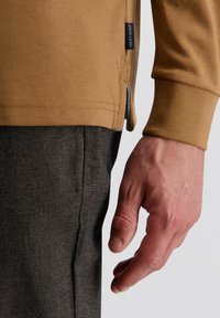 Bruine lange mouwen shirt met een zijsplitdetail, gecombineerd met zwart-wit geblokte broek. Close-up van een hand die tegen de stof rust.