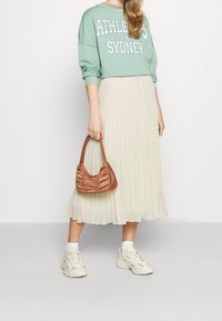 Ljusgrön sweatshirt med vit text, krämfärgad plisserad midi-kjol och liten brun samlad handväska. Vita sneakers med texturerade detaljer.