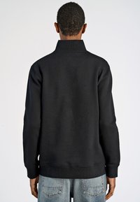 Felpa nera con colletto alto, maniche lunghe e polsini elasticizzati. Il tessuto è liscio con una trama compatta, indossata sopra jeans azzurri chiari.