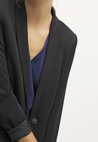 Mustakasta bleiseri, jossa on räätälöity muotoilu, shaalli kaulus, yksitt äinen napitus ja rullatut hihansuut. Päällä tummansinisestä verkkokangasta oleva paita.