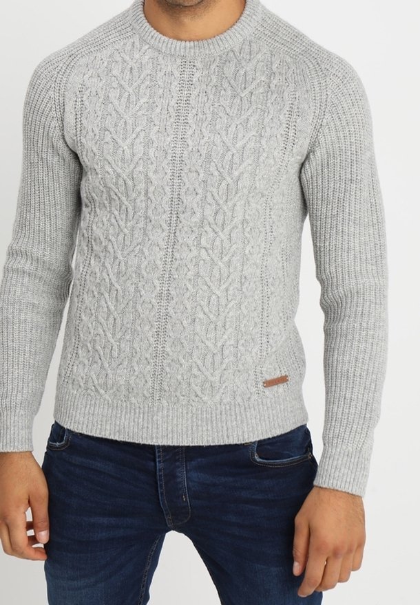 Pull gris en tricot avec un motif en torsades texturé, col rond et poignets côtelés. Porté avec un jean bleu foncé, présente une petite étiquette marron.