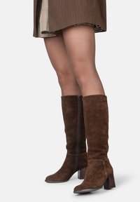 Bottes hautes en daim marron, avec un talon carré, dotées d'une fermeture éclair sur le côté et d'une texture lisse, complétant une jupe marron à rayures.