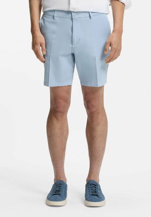 Uomo che indossa pantaloncini su misura azzurri chiari e sneakers blu abbinate, in piedi contro uno sfondo bianco.