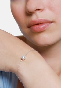 Bracciale d'argento con una singola perla bianca, design in filo sottile, indossato su un polso nudo, su uno sfondo chiaro. Texture morbide visibili sulla pelle.