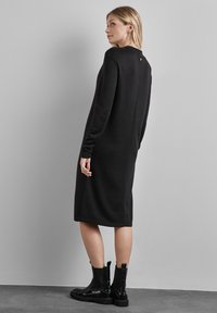 Abito lungo nero a maniche lunghe con scollatura tonda, design elegante e spacco laterale. Indossato con stivaletti neri alla caviglia, dotati di dettaglio con bottone posteriore.