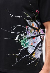 Chemise noire avec un design de surface fissurée révélant des fleurs colorées et des plantes vertes sous les fissures sur le côté.