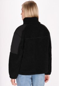 Giacca sherpa nera con colletto alto, maniche larghe e pannelli sulle spalle neri lisci a contrasto. Texture morbida con una vestibilità comoda.