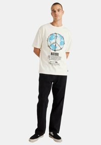 Camiseta blanca con estampado de un globo azul y símbolo de paz, combinada con pantalones negros. Tiene mangas cortas y texto gráfico en la parte inferior.