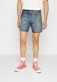 Persona vestida con pantalones cortos de mezclilla de color azul claro con dobladillos deshilachados, calcetines blancos y zapatillas de lona altas de color coral, de pie sobre un suelo gris.