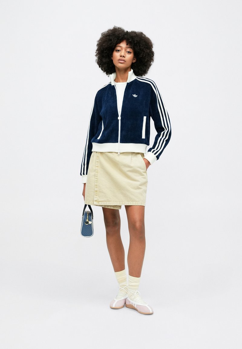 Veste zippée en velours côtelé bleu marine avec des rayures blanches, associée à une jupe beige. Chaussures blanches et roses avec des chaussettes jaunes. Petit sac bleu.