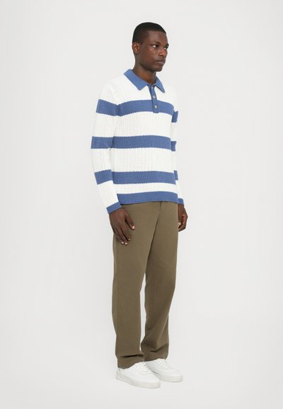 Hvit strikkegenser med blå striper, kort krage og tre knapper, paret med olivengrønne bukser og hvite joggesko.