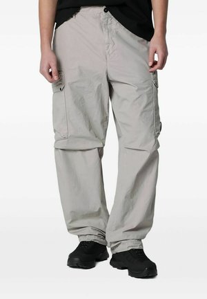 Persona che indossa pantaloni cargo larghi grigio chiaro con tasche con patta e sneaker nere, in piedi davanti a uno sfondo uniforme.