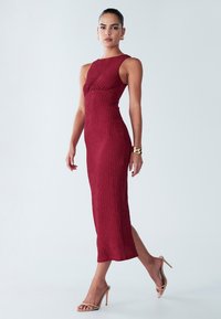 Gojen bordo midi obleka z oprijetim krojem, brezrokavna zasnova in stranski razporek. Oblečena z golo sandali z visokimi petami.