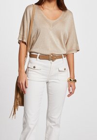 Metalic beige V-hals trui met losse mouwen, gecombineerd met witte strakke jeans met voorzakken en een bruine riem. Eenvoudige gouden armband.