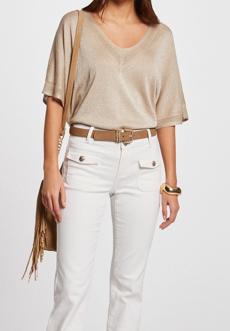 Metalic beige V-hals trui met losse mouwen, gecombineerd met witte strakke jeans met voorzakken en een bruine riem. Eenvoudige gouden armband.