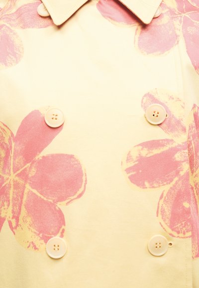 Κίτρινο ύφασμα με μεγάλα ξεθωριασμένα ροζ λουλούδια και τέσσερα μπεζ κουμπιά τοποθετημένα σε τετράγωνο κοντά σε μυτερό γιακά.