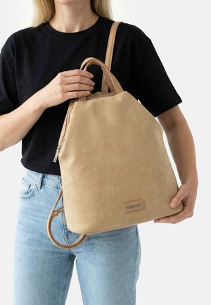 Moteris juoda palaidine ir mėlynais džinsais laiko didelę smėlio spalvos odinę rankinę su viršutinėmis rankenomis ir petnešomis.