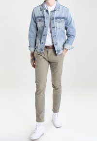 Veste en jean bleue claire, t-shirt blanc, pantalon beige et baskets blanches. Comprend des poches avant et des manches retroussées. Design simple.