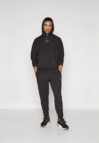 Muž stojí s rukama v kapsách, oblečený v černé mikině s kapucí, odpovídajících joggersech a černých sportovních teniskách na pozadí jednoduché šedé stěny.