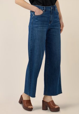 Person trägt dunkelblaue Jeans mit weitem Bein und roher Saumkante sowie braune Plateausandaletten mit offenem Zehenbereich, Hand in der Tasche, vor beigem Hintergrund.