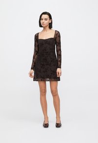 Schwarzes langärmeliges Kleid mit transparentem Überzug und Blumenmuster. Figurbetonte Silhouette mit gerafften Details, oberhalb des Knies lang.