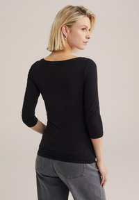 Chemise noire ajustée à manches trois-quarts, décolleté large, et texture douce, associée à un pantalon en denim gris dans un cadre décontracté.