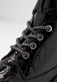 Bottines en cuir verni noir avec une texture crocodile, dotées de lacets tissés noir et blanc et d'œillets en métal argenté.