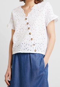 Blouse blanche en dentelle avec un col festonné, des manches courtes et des boutons marron. Portée avec une jupe en denim.