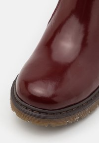 Bottines en cuir bordeaux avec une finition brillante, bout rond, semelle texturée marron et surpiqûres foncées contrastantes le long du bord.